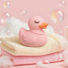 Pink Miss Duck bath-bomb accessory placed on folded towels with bubbles and foam on a soft pink background.