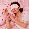 Glampton cherry-print satin sleep set displayed in use, showing the matching satin pillowcase, eye mask and scrunchie in the cherry pattern.